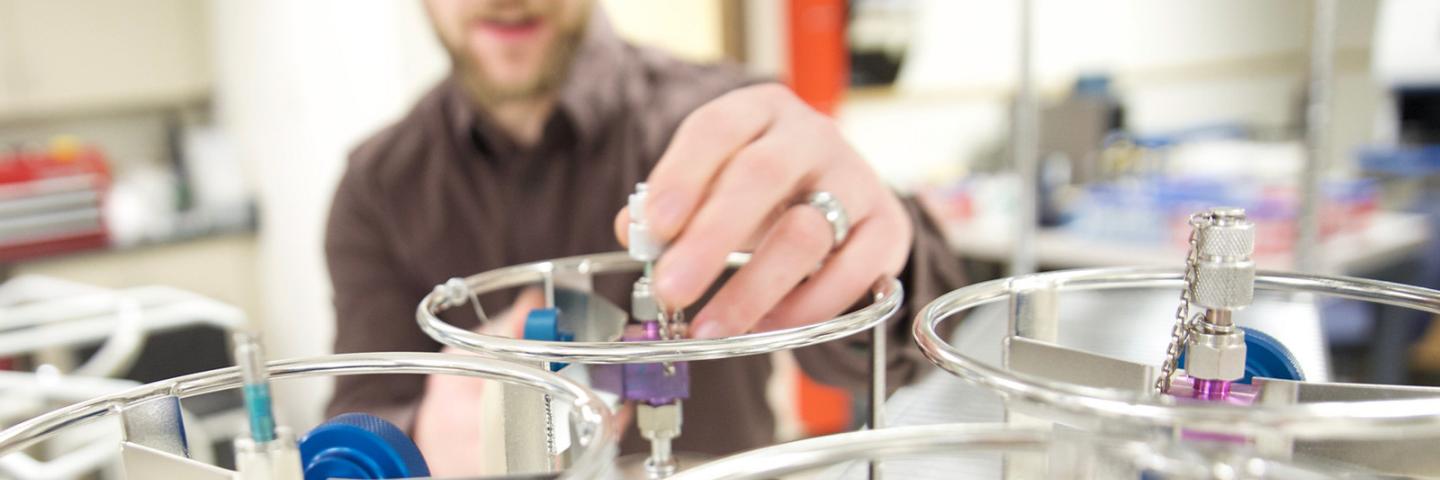 A man in a lab turns a knob on top of a metallic cannister.