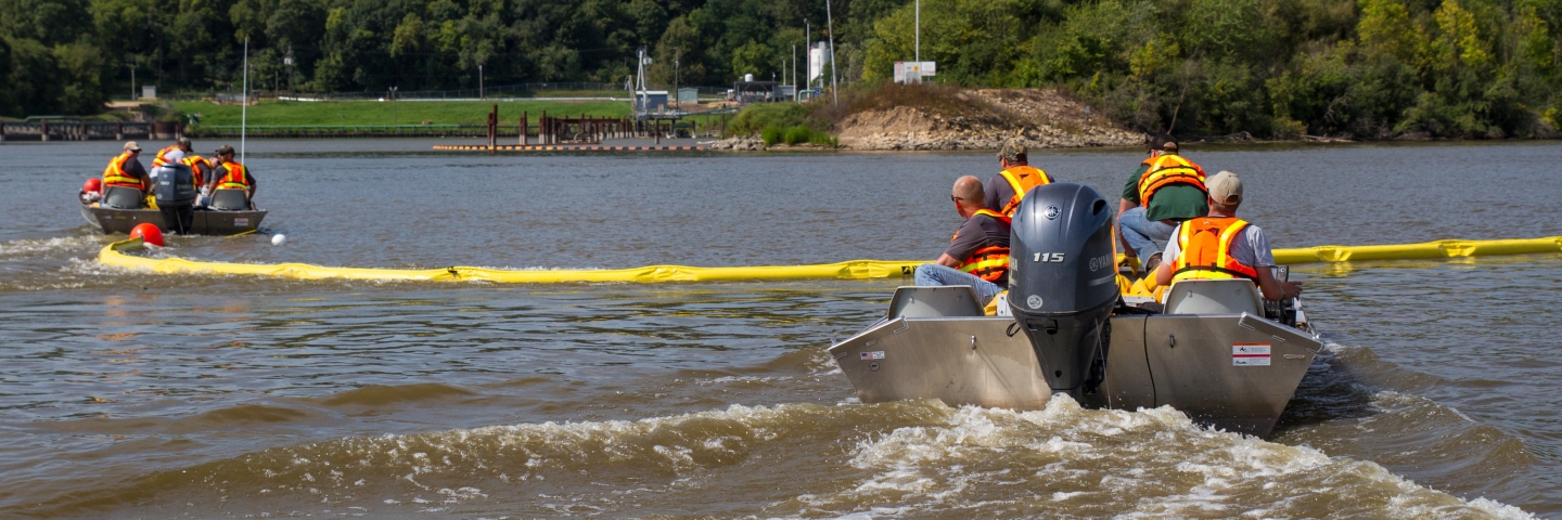 Training exercise for an emergency spill. Two boats on the river pulling a bright yellow plastic spill containment boom between them. 