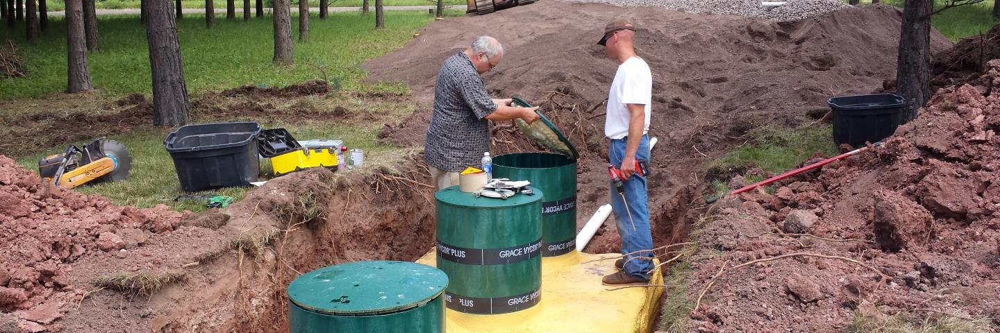 Two men working on a yellow septic system tank in a large trench in the ground. 