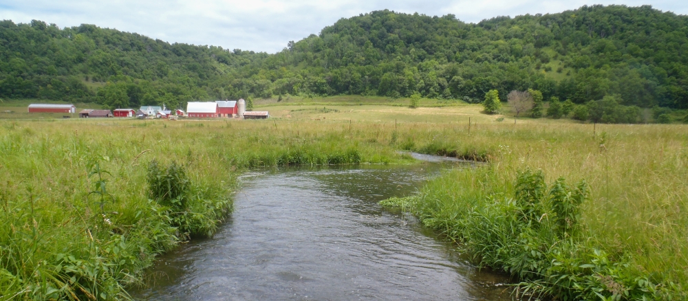 Stream running through grassy field with red barn and farm buildings in background.