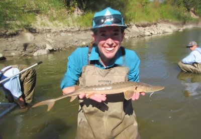 Smiling woman wearing waders standing in a shallow river holding a small sturgeon. 