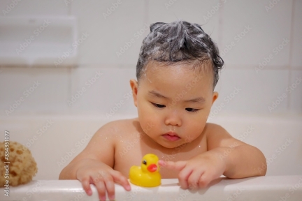 Young Asian girl playing with yellow rubber duck on edge of bathtub.