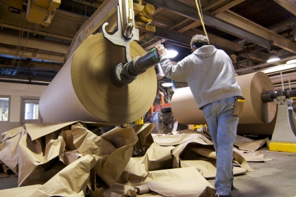 Man standing by large roll of new brown paper produced from recycled paper and cardboard. 
