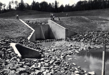 Black and white photo of men standing on a rise with a small concrete dam and culvert.