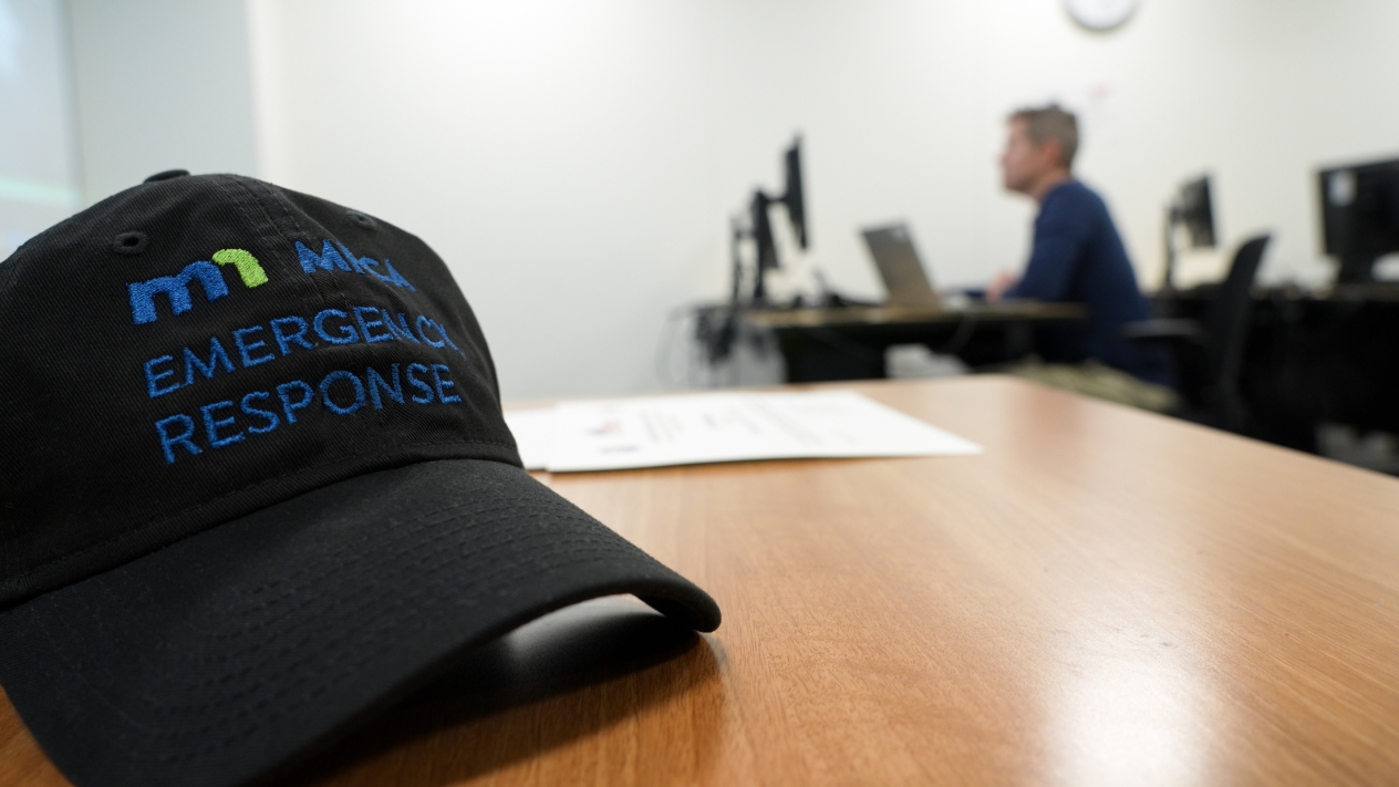 A black hat that reads MPCA emergency response sits on a table. There is a man on a computer in the background.