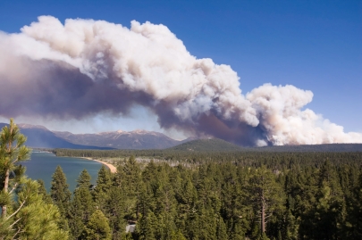 Heavy plume of white smoke rises from pine covered mountains in distance
