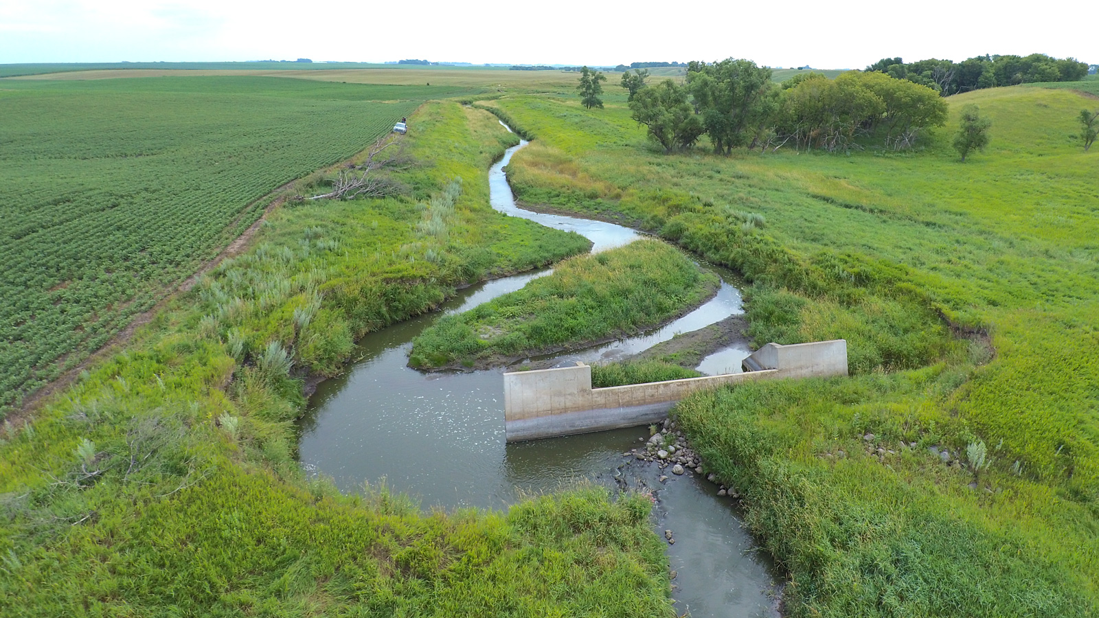 A small stream next to a farm field flows around one side of a concrete dam
