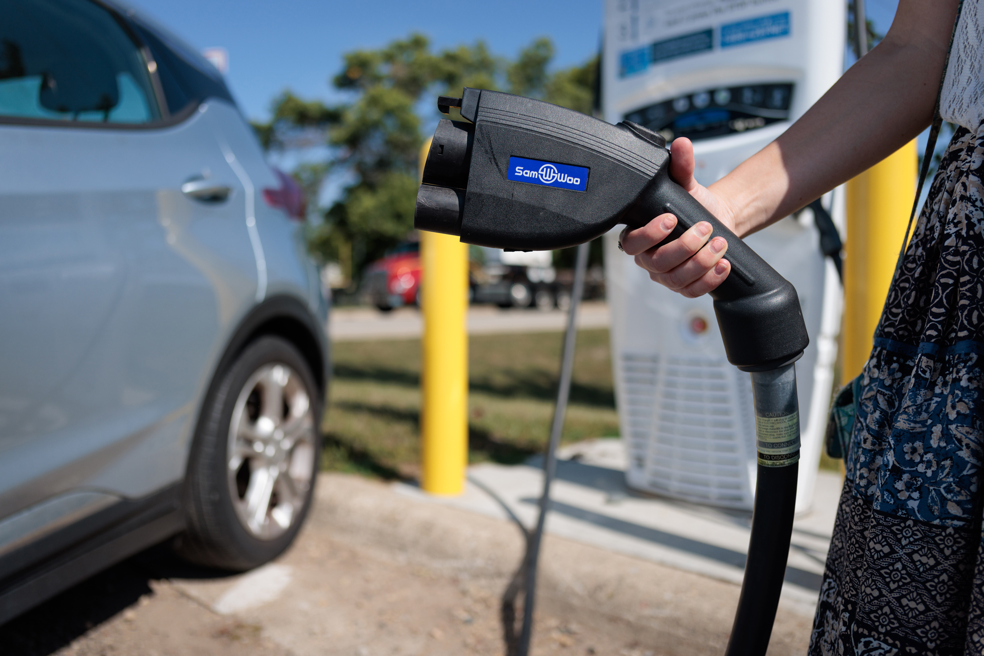 Person holding an EV charging connector. A car and EV charging station are in the background.