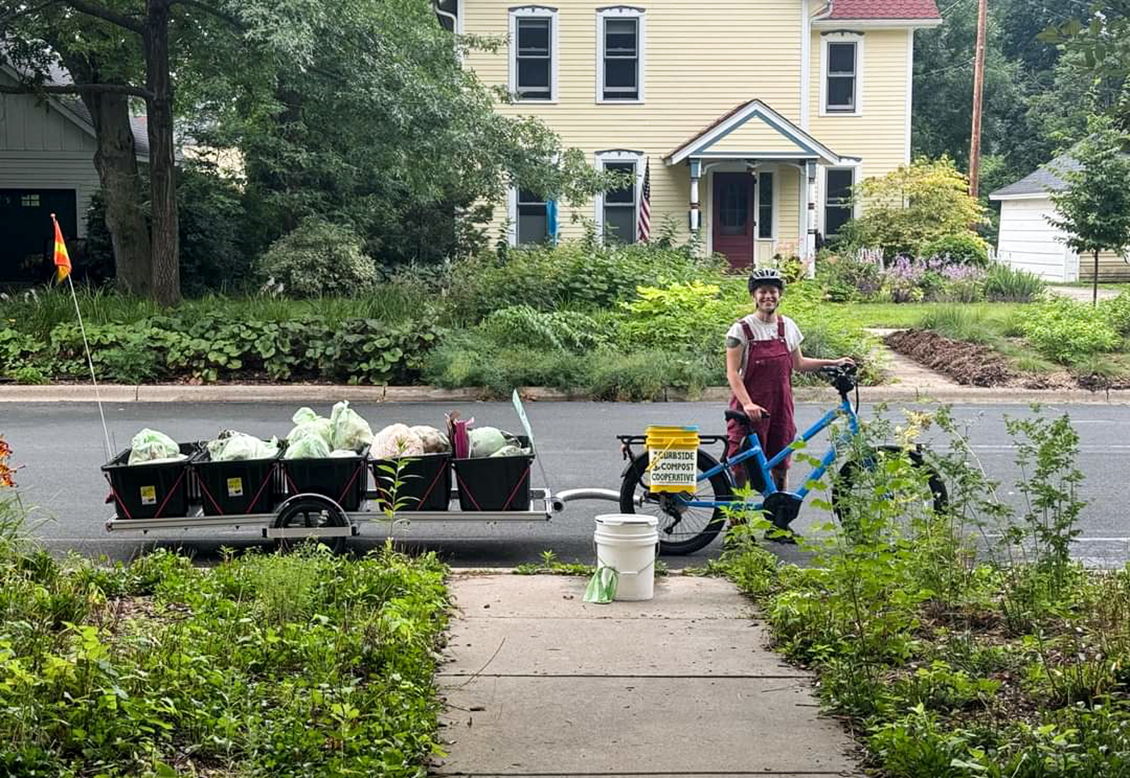 A person standing next to a bike pulling a trailer carrying black bins filled with bags. A bucket on the bike reads "Curbside Compost Cooperative."