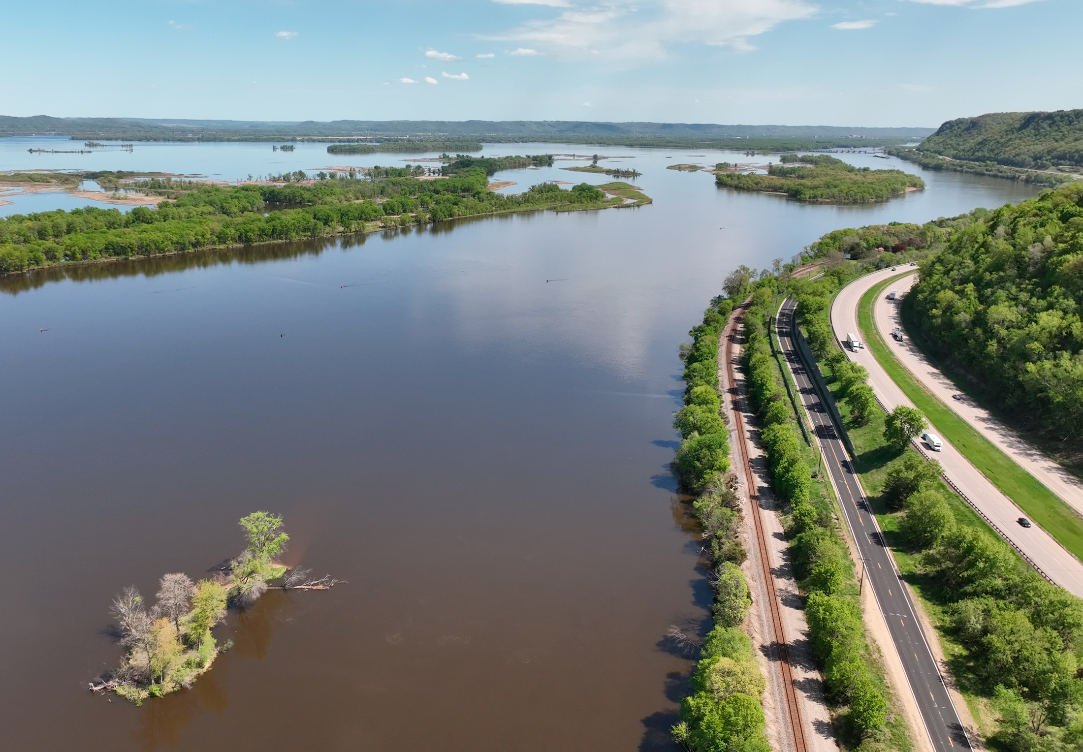 Aerial view of a large brown river with highways and railroads tracks running along the banks on the right side.
