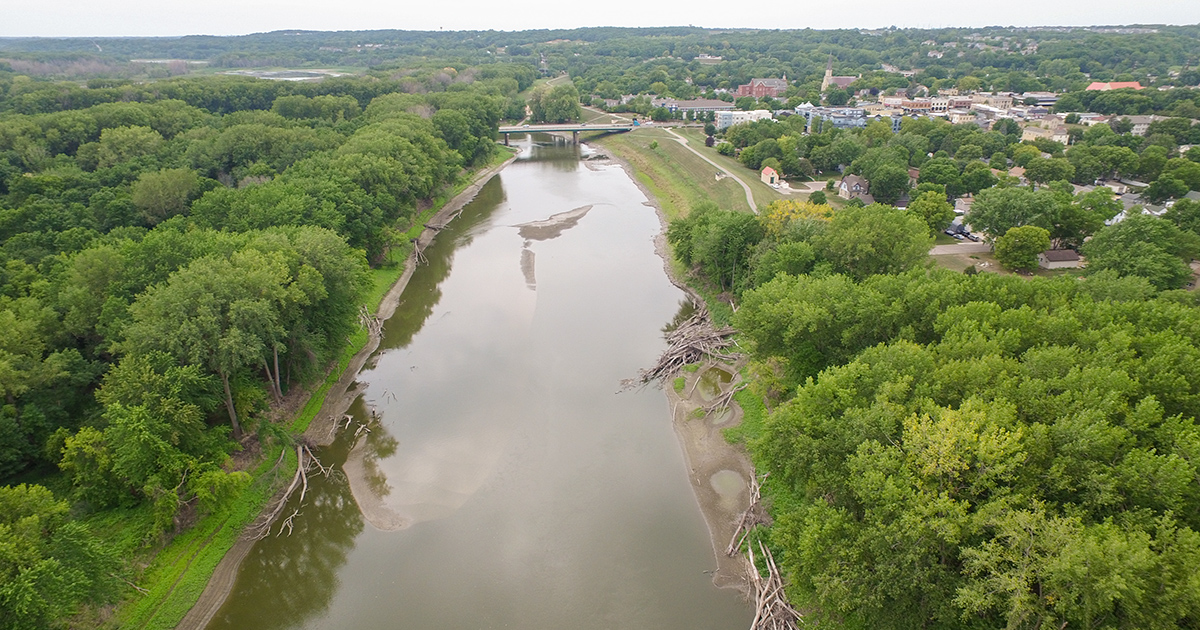 Aerial view of a brown river with tree-lined banks, a town on the right side, and a bridge in the distance.