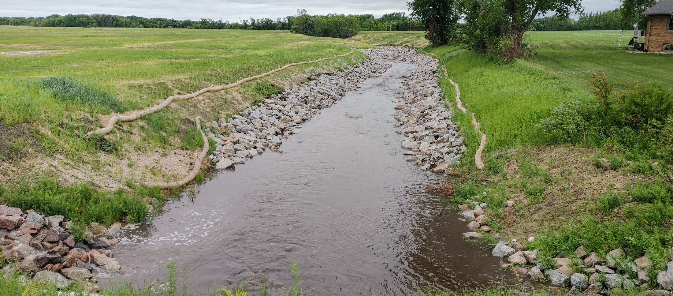 Water flowing through a shallow ditch with rock stabilized banks to prevent erosion. 