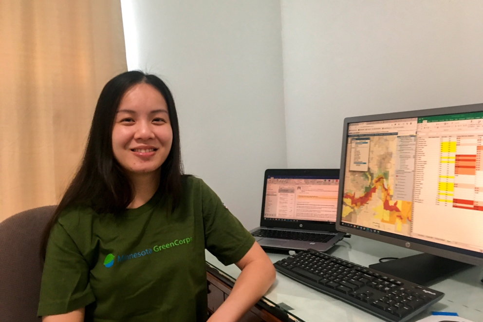 A smiling woman wearing a Minnesota GreenCorps t-shirt sitting at a desk in front of a computer. 