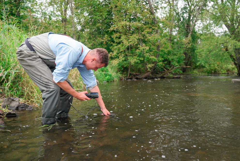 A person leans down to collect a sample for biomonitoring, testing water for temp, clarity and measuring stream width.