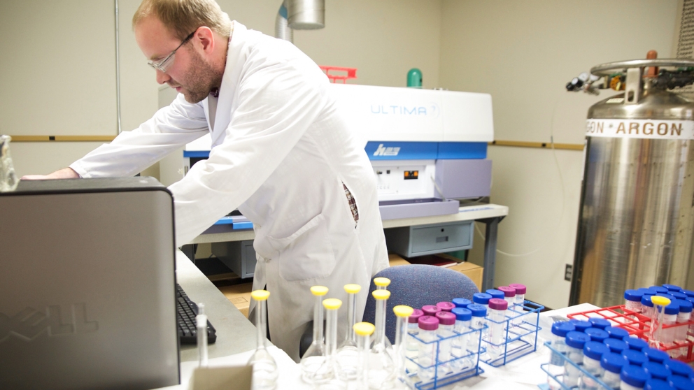 A man in a white lab coat looks at a computer. Flasks and tubes are in stands on the surface next to him.