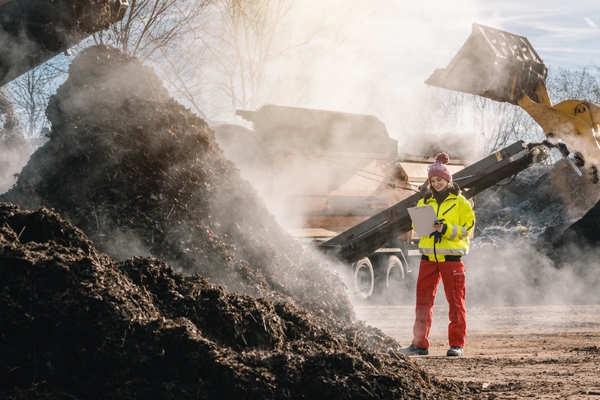 Woman with clipboard standing next to pile of steaming compost in commercial compost facility.