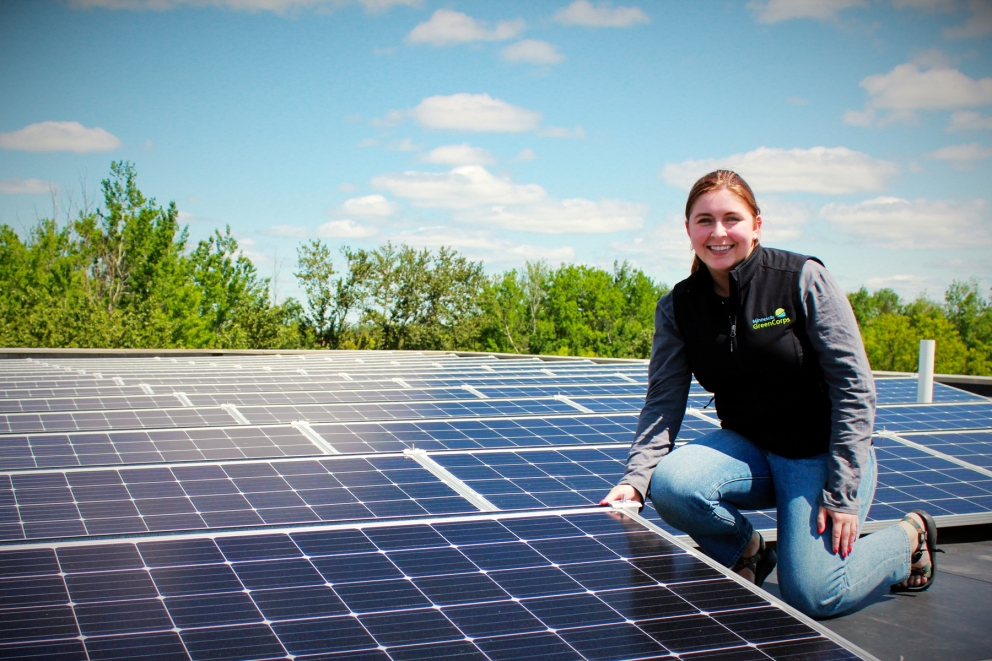 Smiling woman wearing GreenCorps vest kneels next to solar panels.