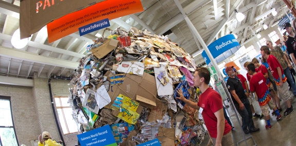 A man looking up at a large ball made of paper products that were thrown away instead of recycled.
