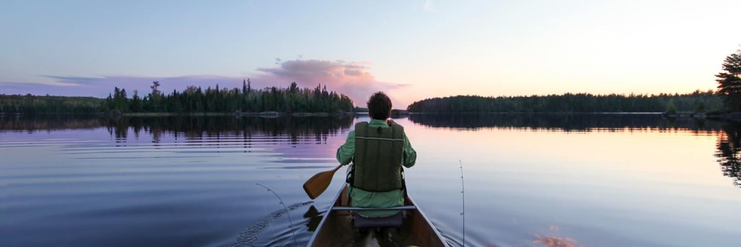 Man with paddle sitting in the front of a canoe with pink and purple clouds reflected in calm surface of the lake.