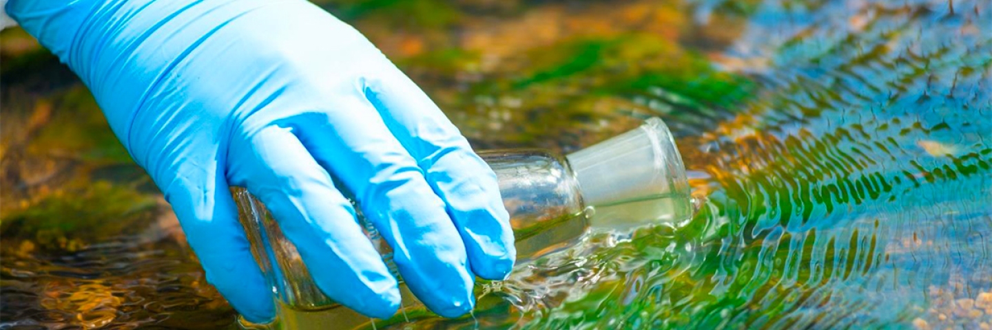 A hand wearing a blue glove dips a bottle in water to collect a sample