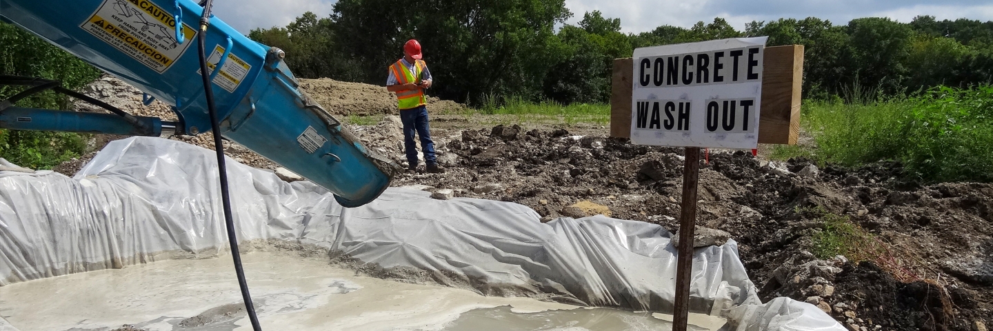 Construction worker stands by plastic lined pit filled with wash water from a concrete mixer and a sign that says "concrete wash out". 