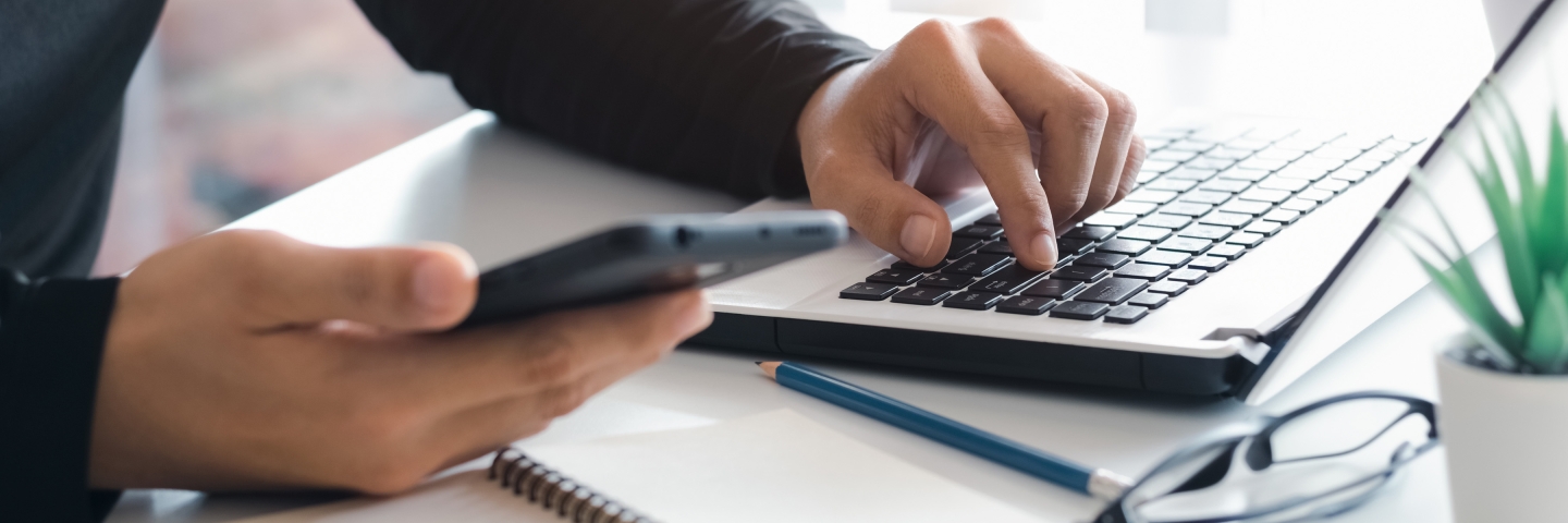 Close up of one hand holding a cell phone, the other hand on a laptop keyboard.