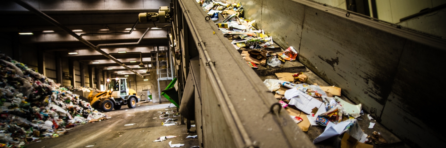 Interior of a recycling facility. A conveyor belt with waste is in the foreground, piles of waste and a bulldozer are in the background.