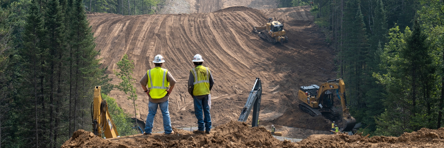 Two construction workers look over cleared space through a forest.