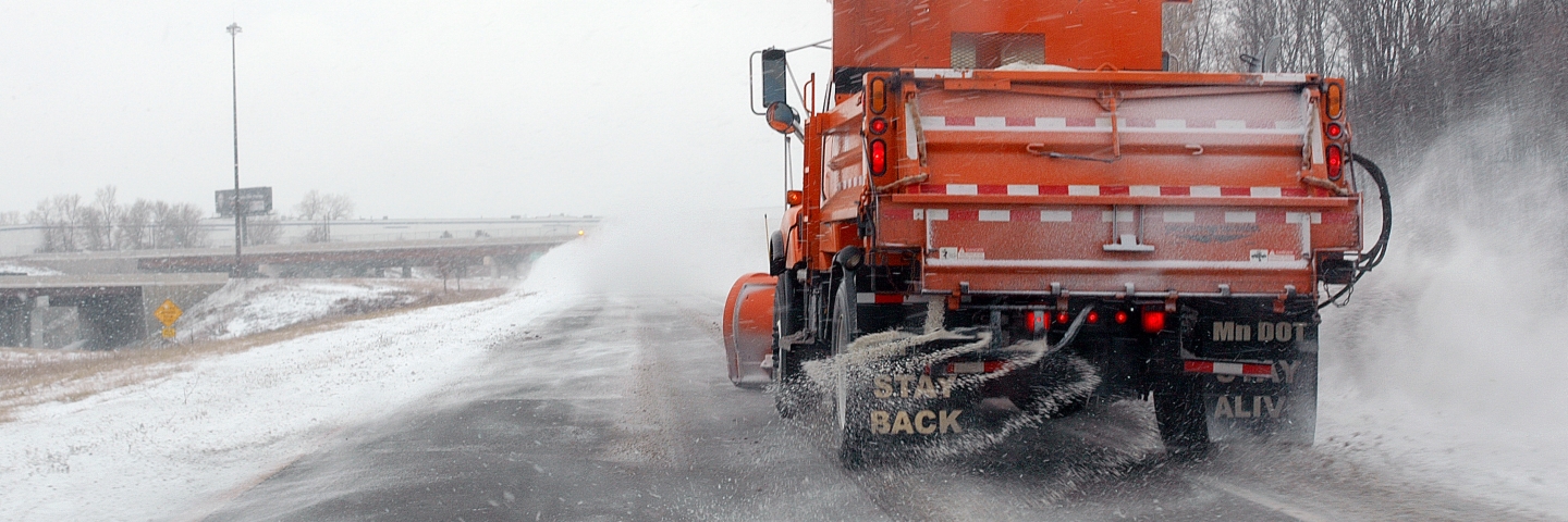 Rear view of an orange snow plow dropping salt on a snowy road.
