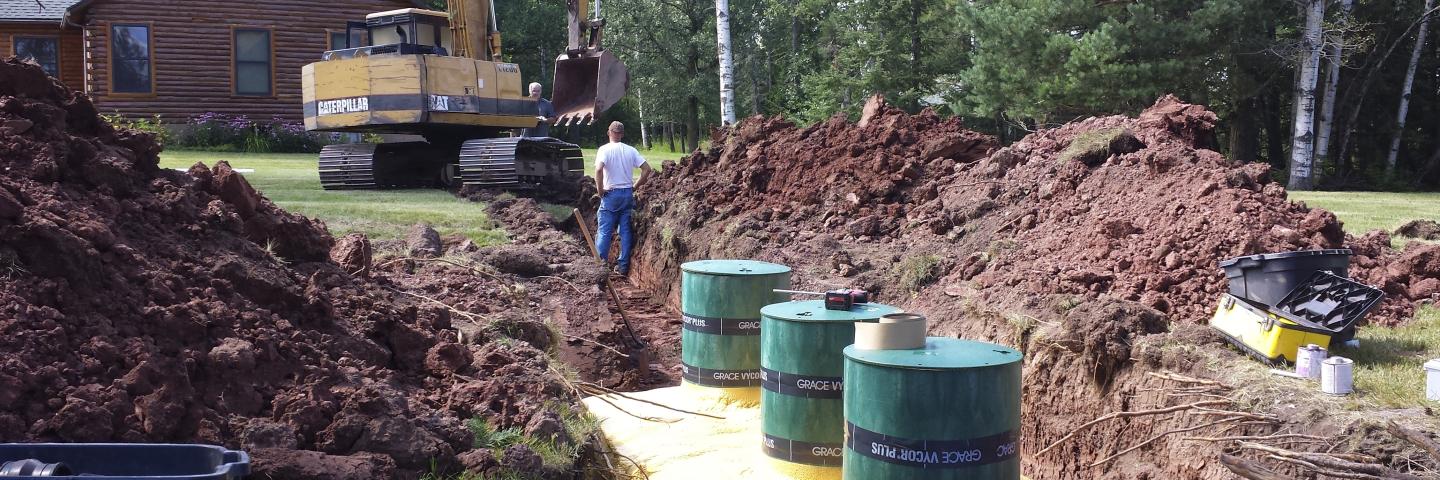 Excavator, mounds of dirt, and a yellow and green septic tank in the backyard of a cabin.