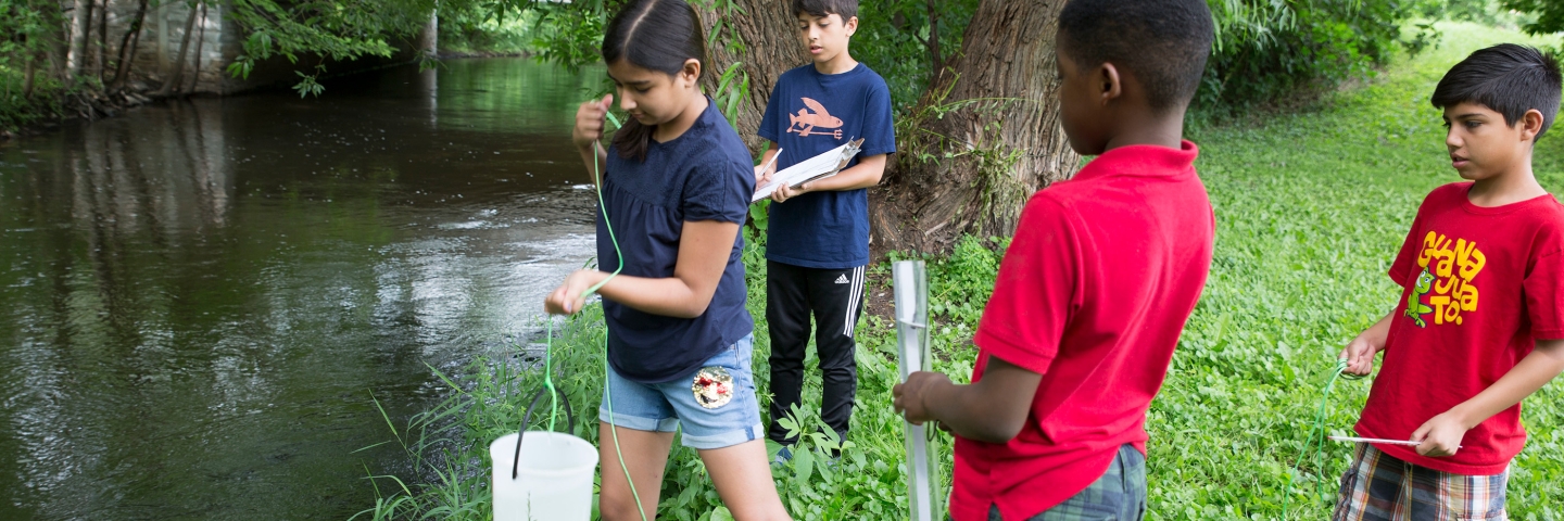 Four middle school children use a bucket and a Secchi tube to collect stream monitoring data.