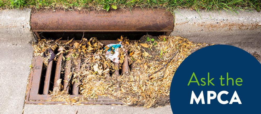 Dried leaves and other debris cover up half of a storm drain grate in a street.