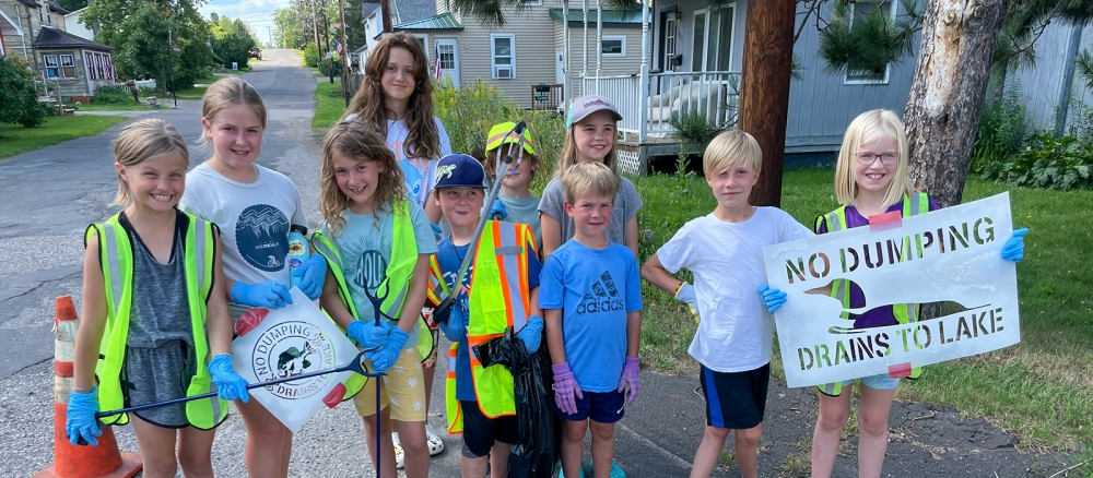 Group of children standing next to a storm drain where they've stenciled "No dumping, drains to lake"