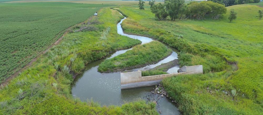A small stream next to a farm field flows around one side of a concrete dam