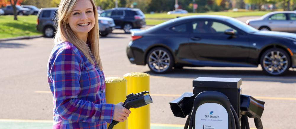 A smiling woman standing next to a electric vehicle charging station, holding the charging cord