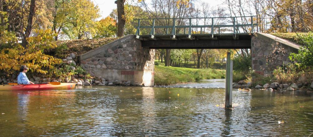 A person in a red and orange kayak floats down a calm stream, about to pass under a small foot bridge. The leaves on the surrounding trees are yellow and green.