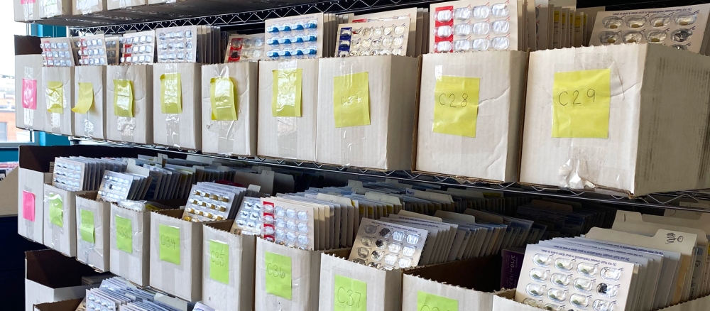 Open top boxes on wire shelves filled with blister packs of prescription medications.