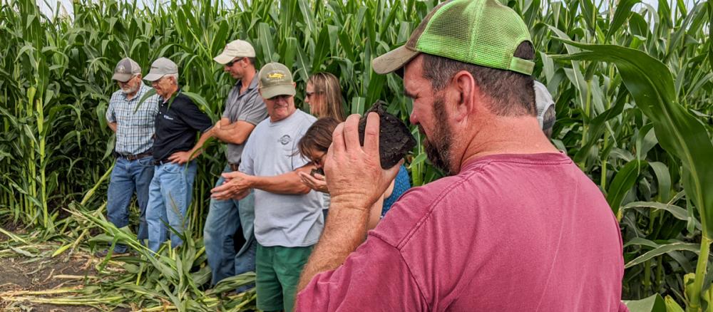 A group of people stand in a clearing in a corn field, a man in the foreground wearing a red t-shirt and lime green cap smells a large clump of soil.