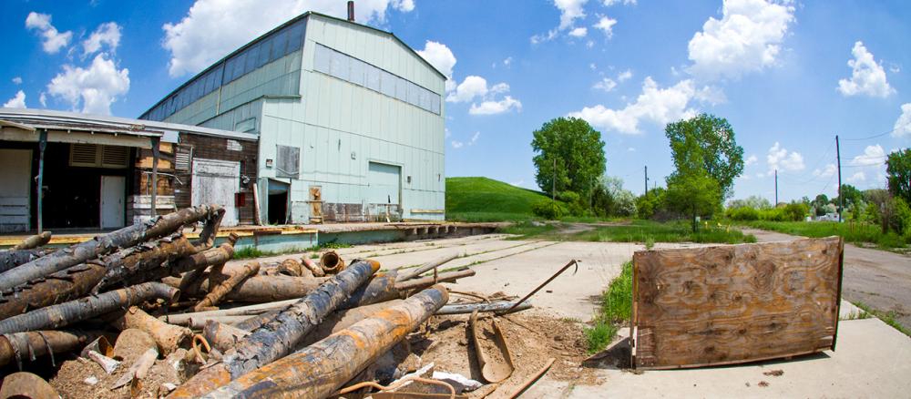A pile of rusted black metal pipes on the ground with an abandoned corrugated metal building in the background