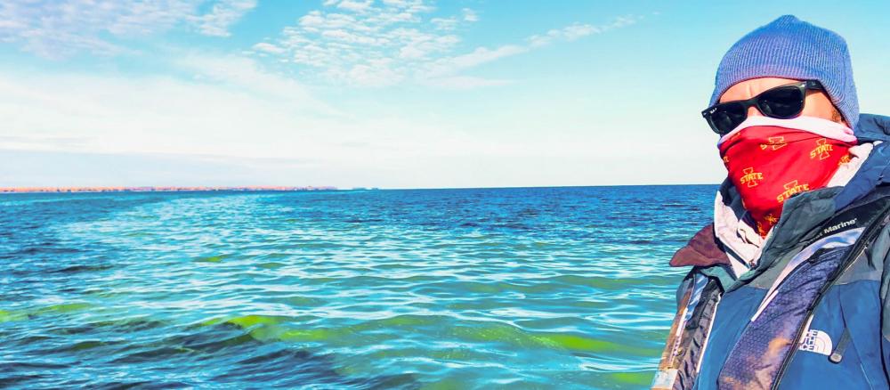 Man wearing winter clothes and sunglasses stands in front of large lake with a wide streak of bright green algae that runs to the horizon.