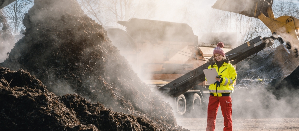 Woman with clipboard standing next to pile of steaming compost in commercial compost facility.