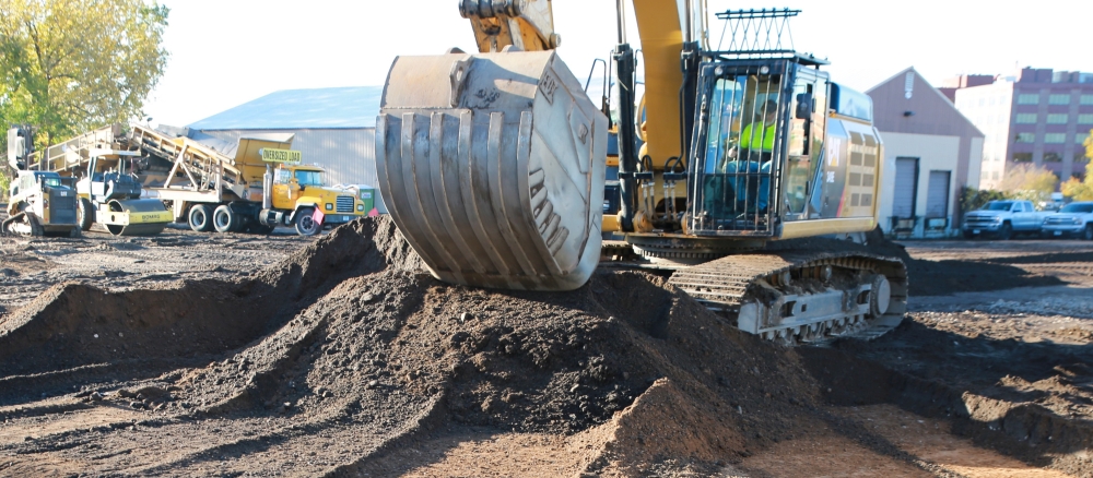 An excavator in an urban environment moving dirt into a pile to expose soil below.