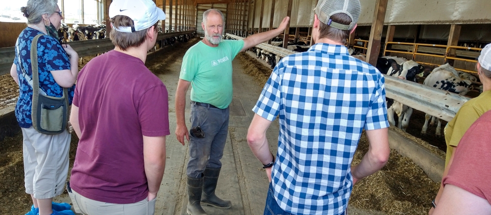 A man speaking to a group of people in a dairy barn with cows in the background.  