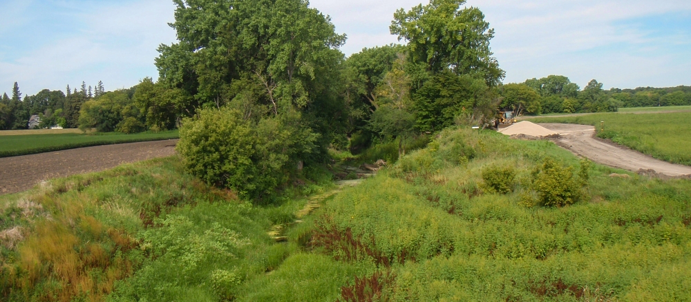 Narrow stream runs through banks overgrown with grass and trees next to a farm field.