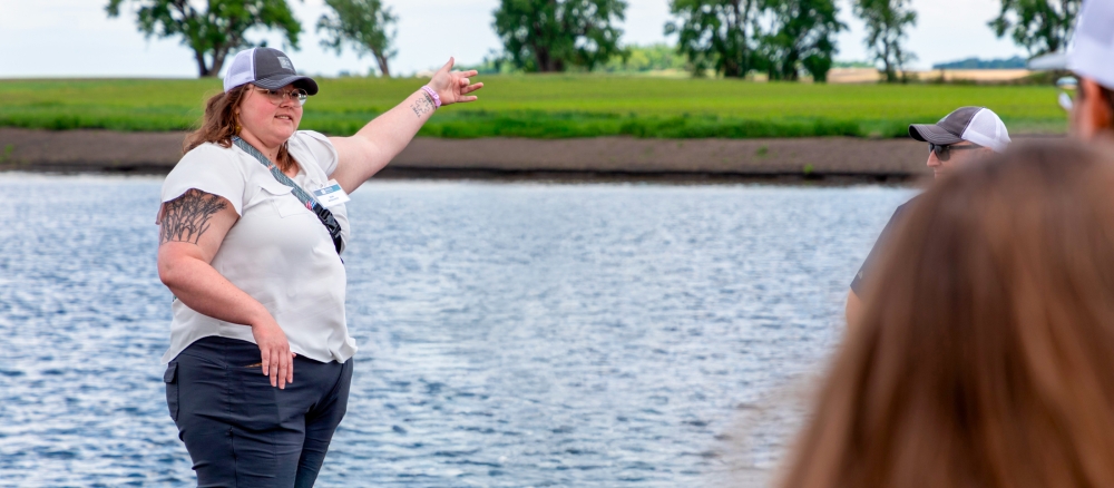 A woman wearing glasses and a cap stands next to water and talks to a group of people.