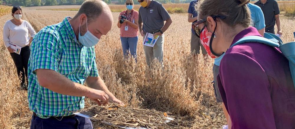 A man in a blue and green plaid shirt holds a white board with a pile of dried vegetation to explain the importance of crop residue to a group of people standing in a farm field.