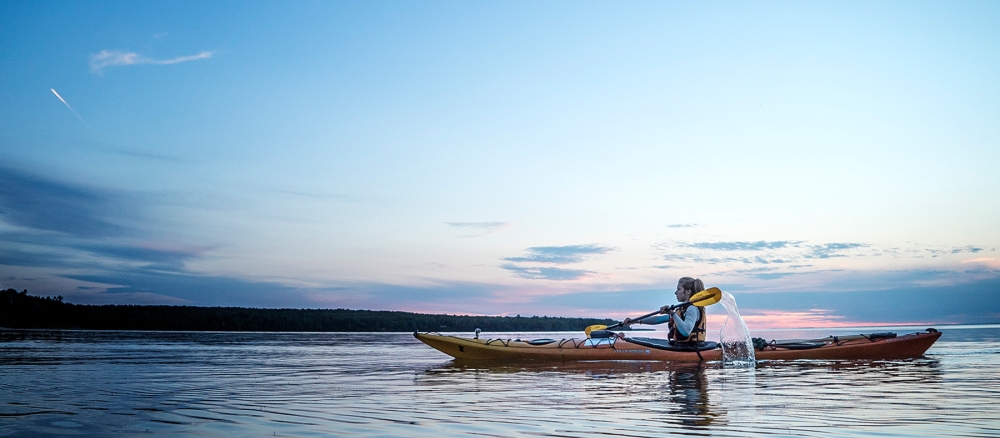 Woman kayaks across a calm lake at sunset.