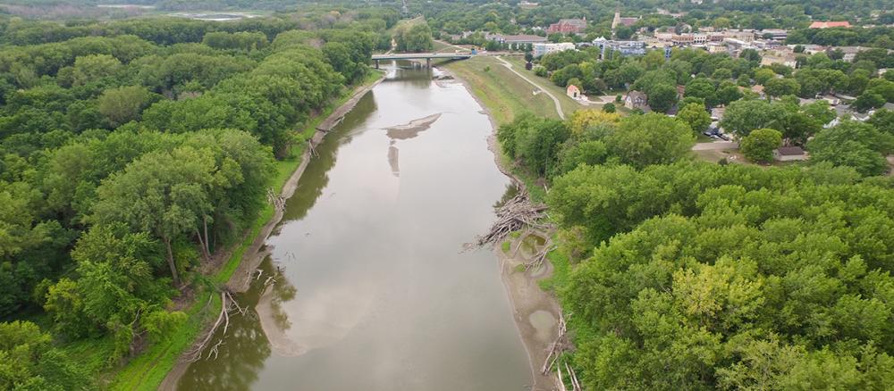 Aerial view of a brown river with tree-lined banks, a town on the right side, and a bridge in the distance.