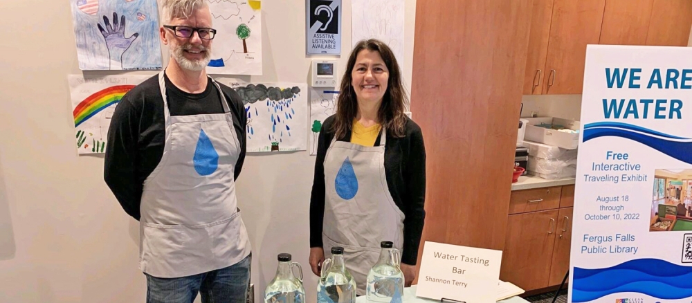 A man and a woman stand behind a table with 3 bottles of water that are used for taste testing samples from different areas of Otter Tail County.