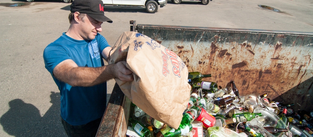 A man emptying a bag of cans and bottles into a recycling dumpster.
